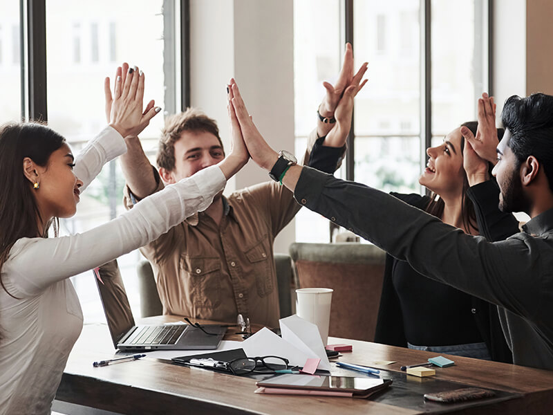 A group of four collogues high-fiving each other around a table. They are in a modern office setting. 