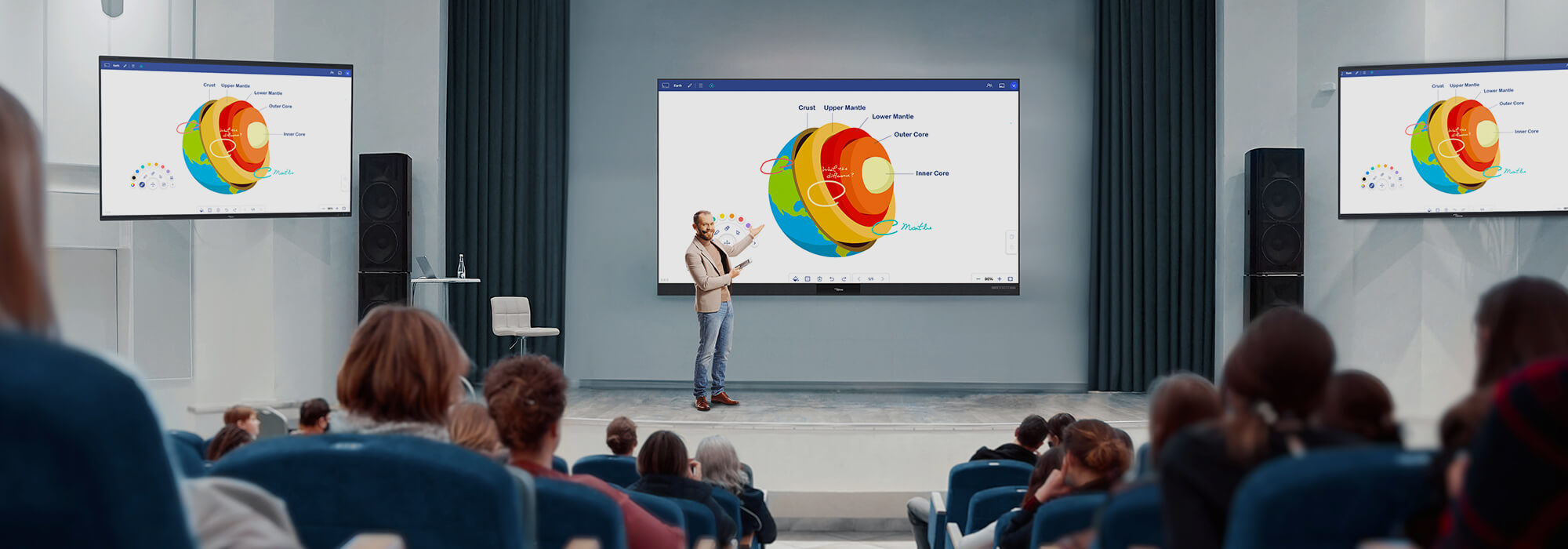 A modern conference room equipped with multiple large interactive displays showing detailed Earth science diagrams. A presenter engages with the audience while demonstrating geological layers and planetary structures on the central screen, with additional displays on either side providing supplementary visual information. The professional setup enables dynamic, educational presentations for corporate training or academic lectures.