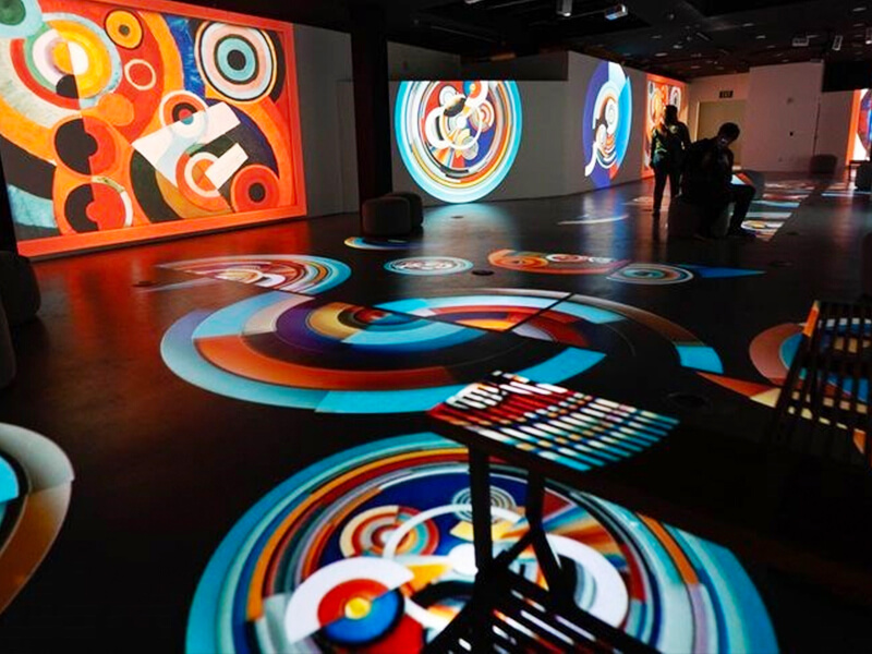 A dark room with multiple screens and projectors. Across the floor, the screens and the walls are digital displays. These are colourful swirls, circles and other patterns. The colours are primary and secondary colours and are bright.