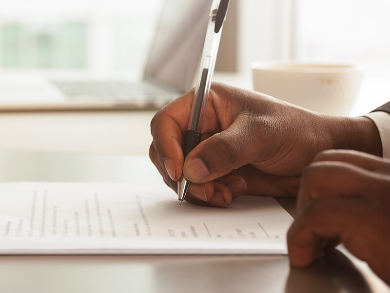 paperwork on a desk and a close up of two sets of hands, each person holding a pen. 