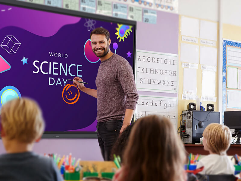 Male teacher in front of a purple Optoma digital flat screen which has the words 'World Science Day' with a smiley face written on it. He is stood in front of a classroom, we can see the back of children's heads. 