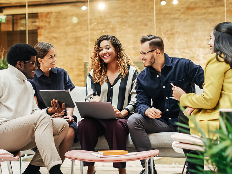 A group of people in a huddle space with laptops and a tablet. They are in discussion and smiling. 