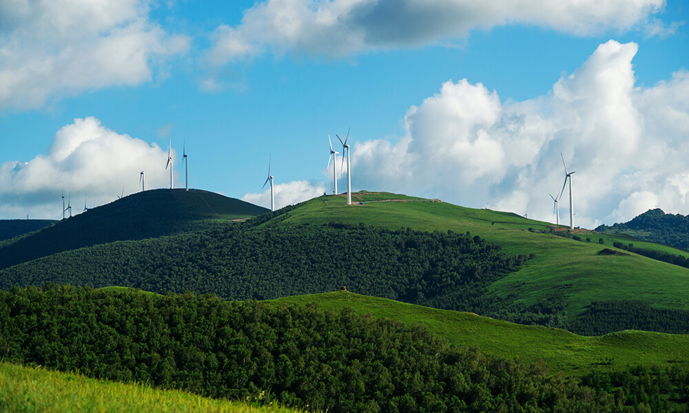 A light blue sky with some white fluffy clouds. Below this are beautiful green hills and at the top of the hills are wind turbines.