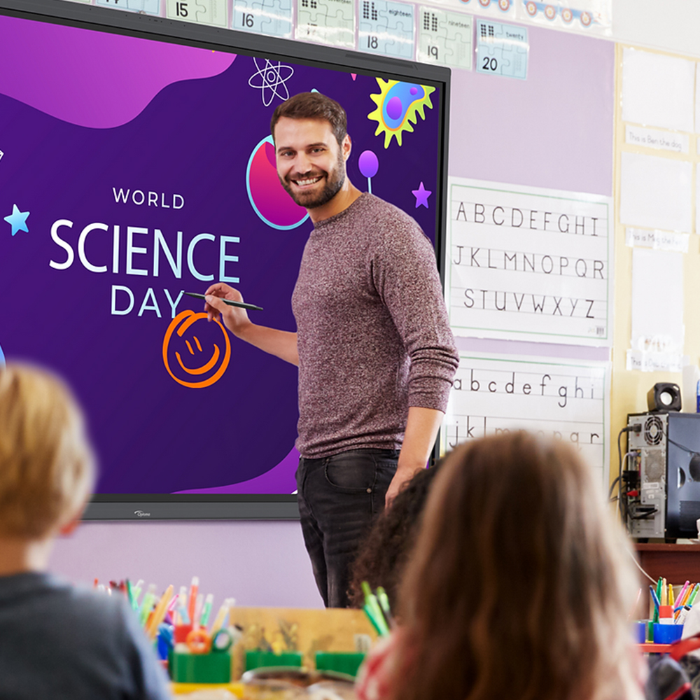 There is a male teacher looking towards the classroom he is teaching. He is wearing a beige cotton jumper with the sleeves rolled up. Behind him is a purple flat panel screen with the words 'world science day'. The teacher is smiling.