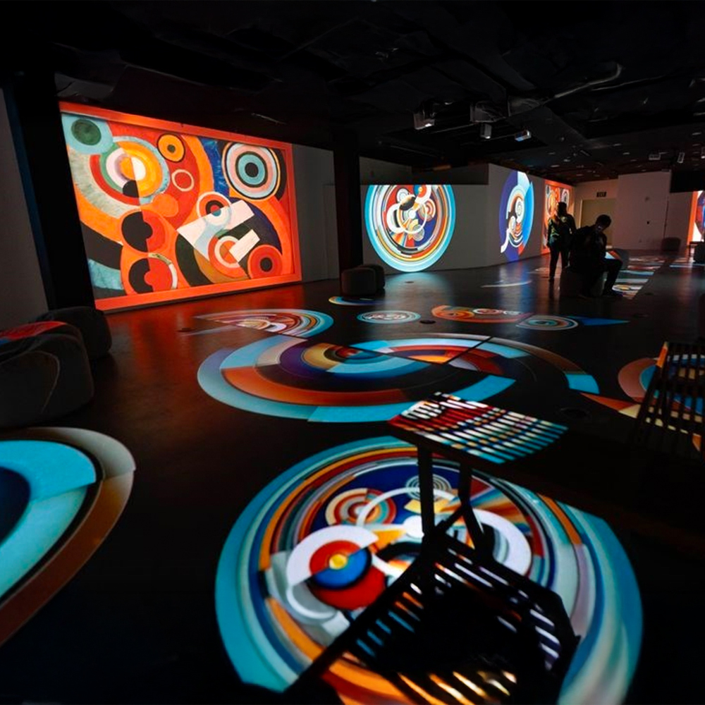 It's a dark room with multiple screens and projectors. Across the floor, the screens and the walls are digital displays. These are colourful swirls, circles and other patterns. The colours are primary and secondary colours and are bright.