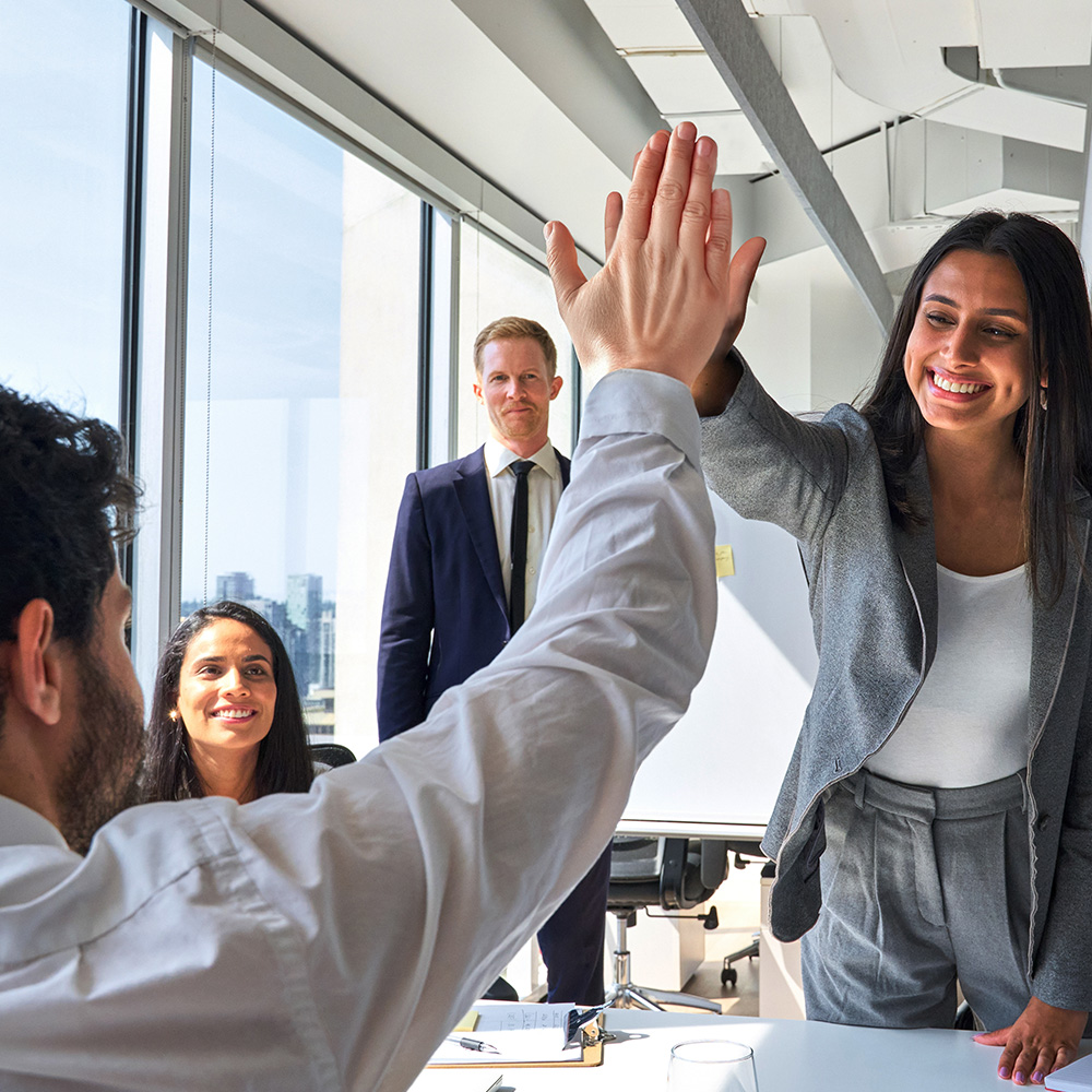 Modern office space with a group of co-working smiling and high-five in front of a large window offering great natural light.
