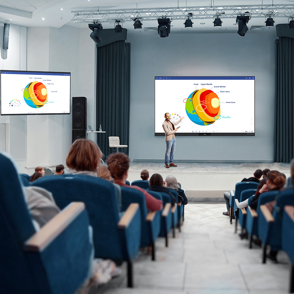 The setting is a lecture room. We can see the back of students sitting in chairs to the left and right of the image. In front of the room is a lecturer  who is wearing a cream blazer and jeans. He has short cut hair and has facial hair. He is in from of a digital flat panel and on the screen is an image of the globe and the various layers within it. To the left of the image we can see a second screen, which has the same globe on it.