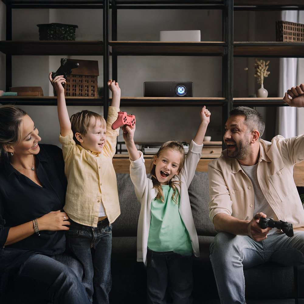 The setting is a front room, the image is of a family on the sofa. From left to right: The mother who is looking lovingly at her family, she has dark blonde hair and is wearing a black blouse. Infront of her is a young boy who has his arms in the air and he is holding a game control, as he is cheering. To his right  is a eight year old girl who has her arms in teh air and is holding a game control also. She is cheering. Her dad is to her right, he is sitting down on teh sofa and looking at his family as he cheers also. Behind then our shelves with various objects and ornaments. We can clearing see an Optoma projector which is switched on.