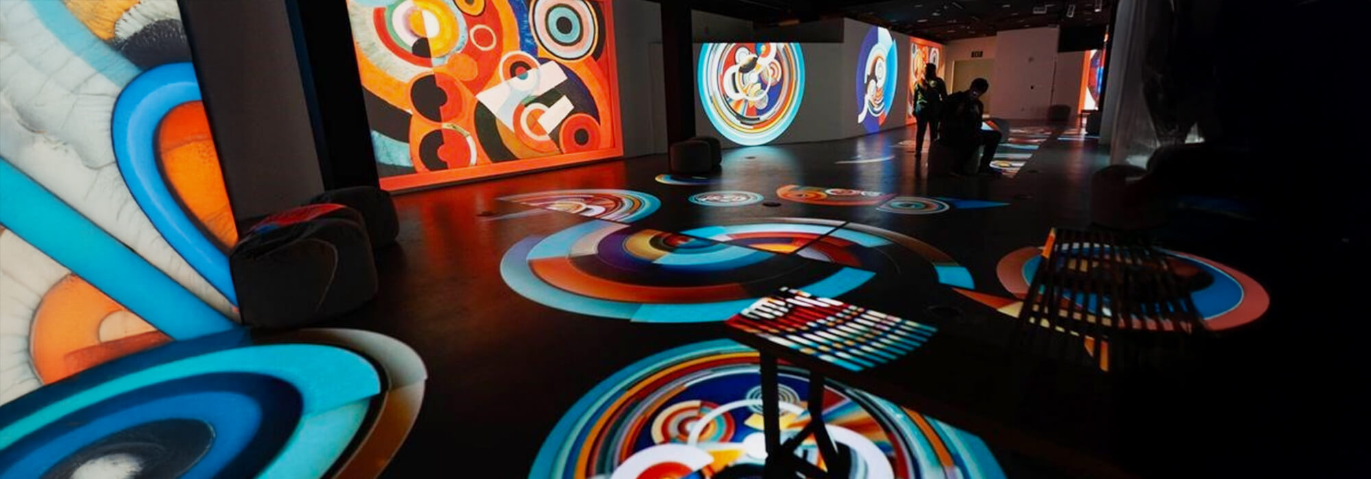 A dark room with multiple screens and projectors. Across the floor, the screens and the walls are digital displays. These are colourful swirls, circles and other patterns. The colours are primary and secondary colours and are bright.