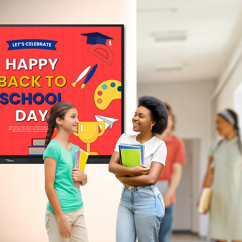 Two female students talking in school hallway near flat screen display showing Happy Back to School Day poster