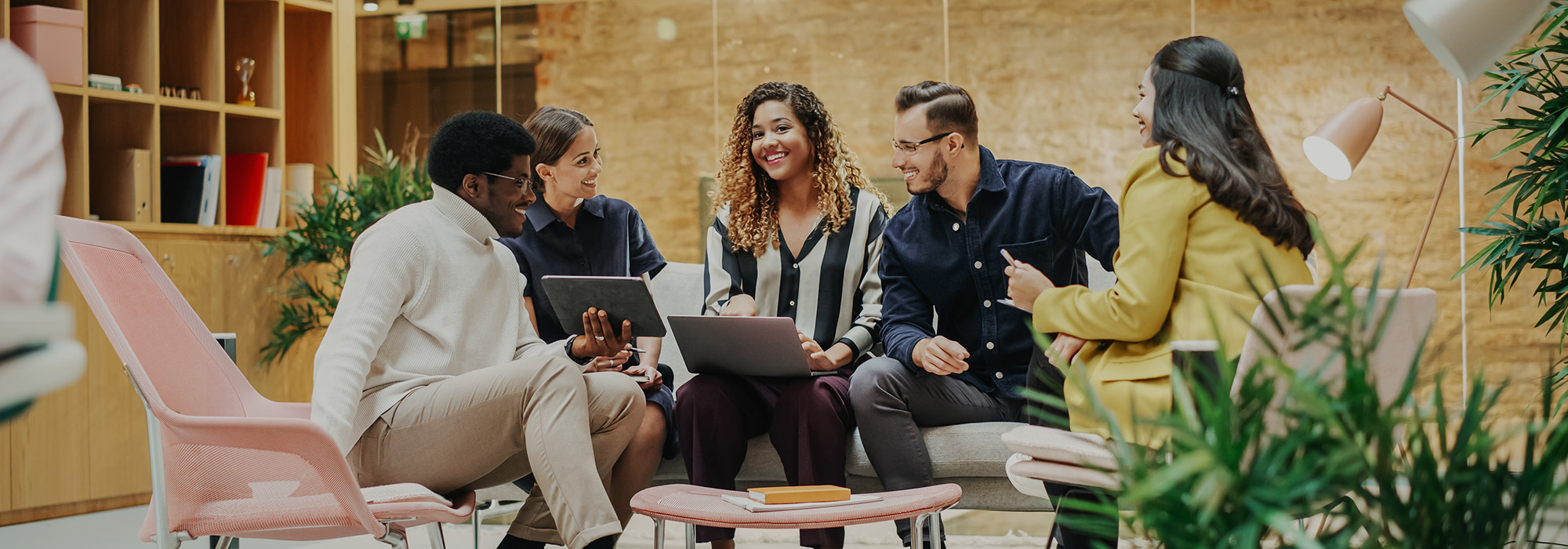 Diverse team of young professionals collaborating with laptops and tablets in modern coworking space with plants and contemporary furniture