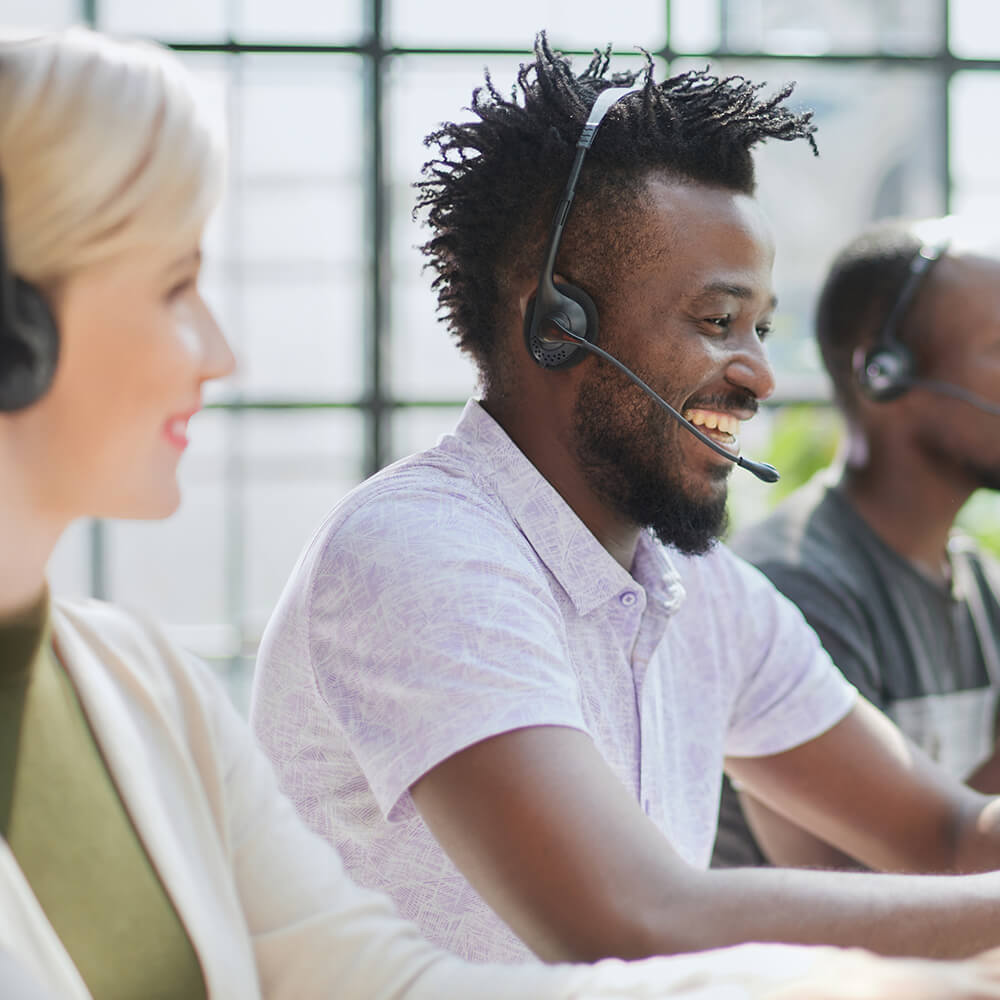 A close up of a man and women at a customer service call centre. The image is focused on the man who has his head-sets on and is smiling as he speaks.