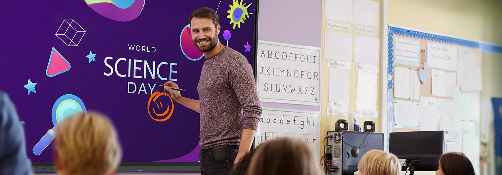 Male teacher in front of a purple Optoma digital flat screen which has the words 'World Science Day' with a smiley face written on it. He is stood in front of a classroom, we can see the back of children's heads. 