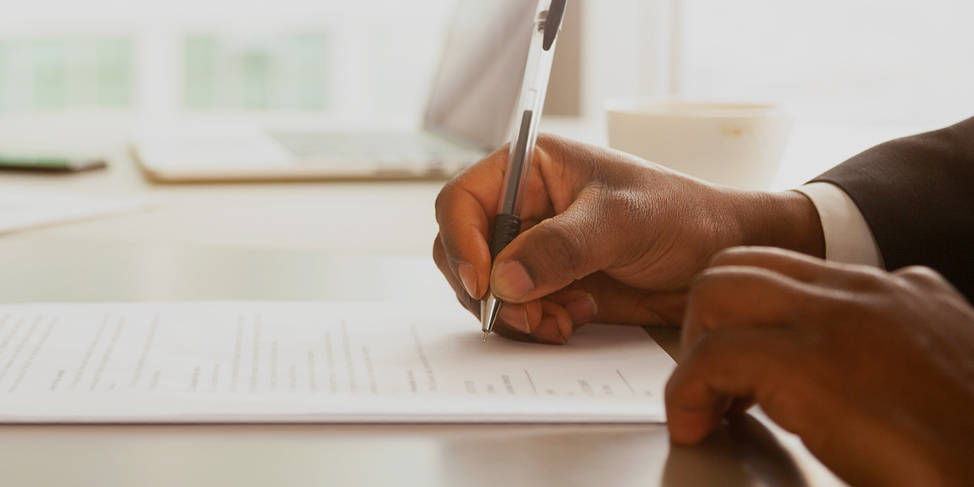 paperwork on a desk and a close up of two sets of hands, each person holding a pen. 