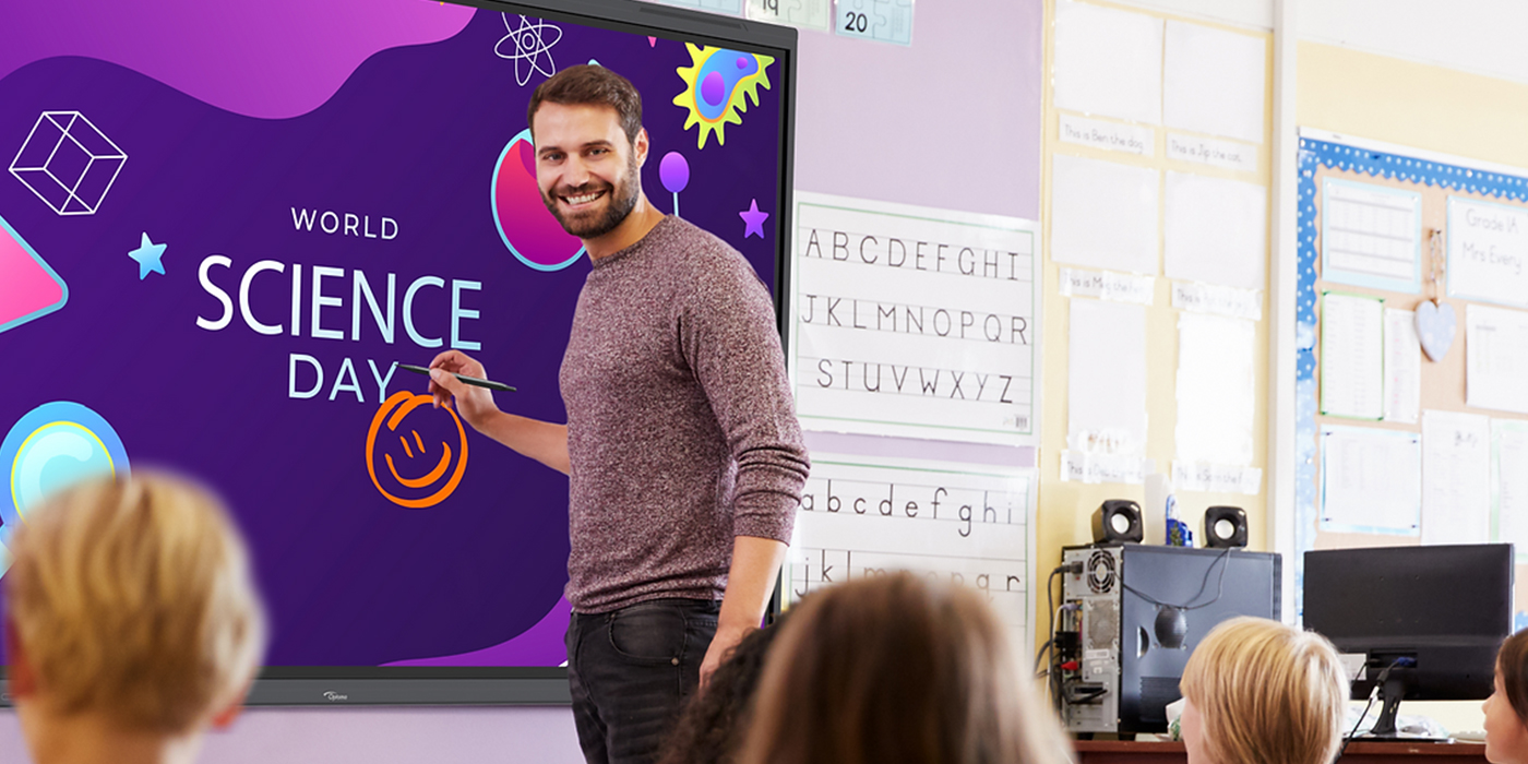 Male teacher in front of a purple Optoma digital flat screen which has the words 'World Science Day' with a smiley face written on it. He is stood in front of a classroom, we can see the back of children's heads. 