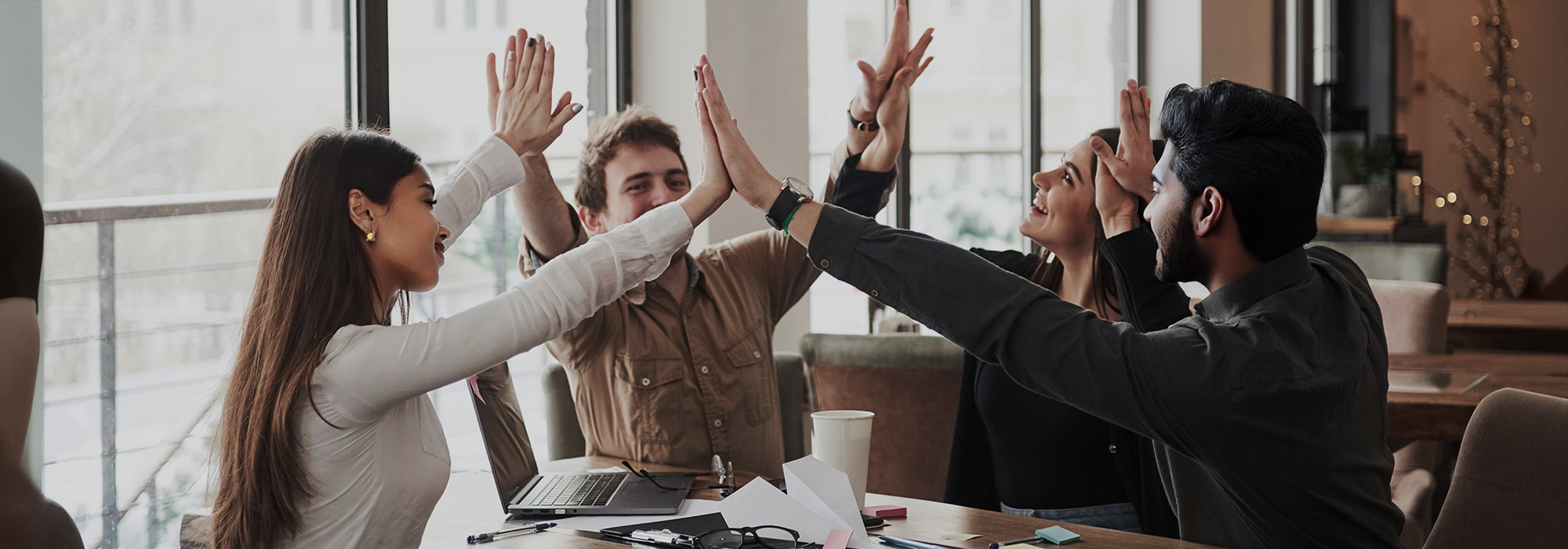 A group of four collogues high-fiving each other around a table. They are in a modern office setting. 